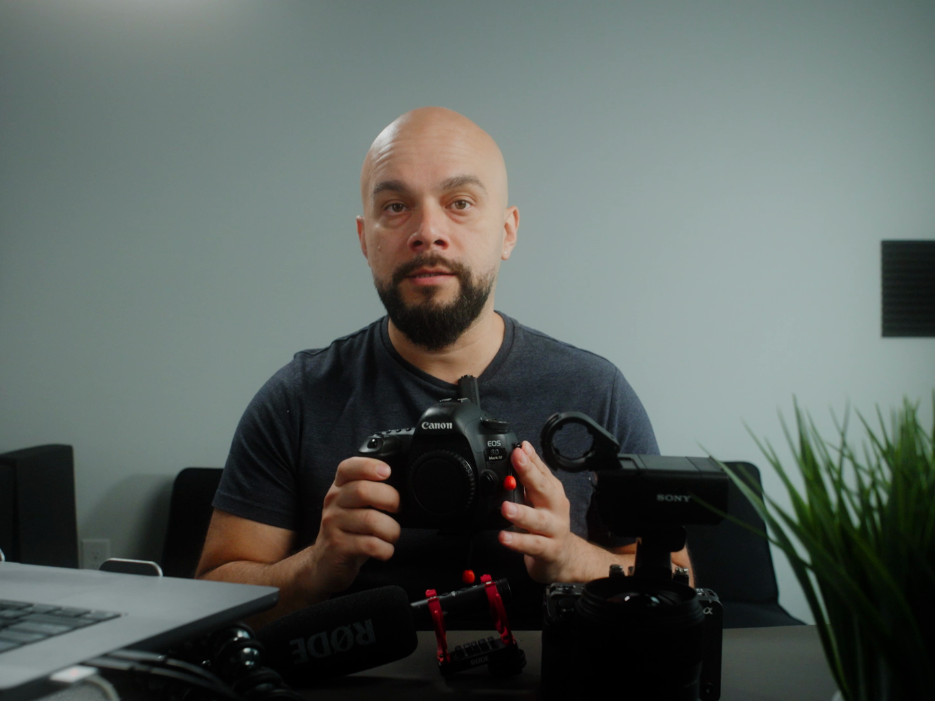 Oscar at his desk holding a Canon camera body, with a Sony body, a Rode microphone and a rig on the desk in front of him.